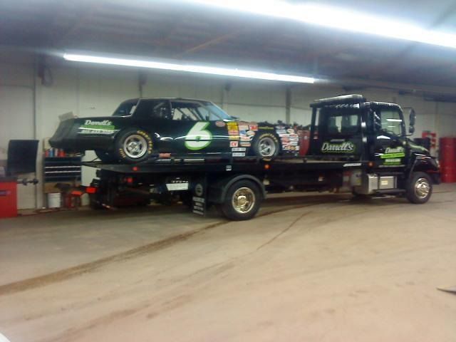 Black race car on a black tow truck inside a garage. The truck has gold rims and lettering.