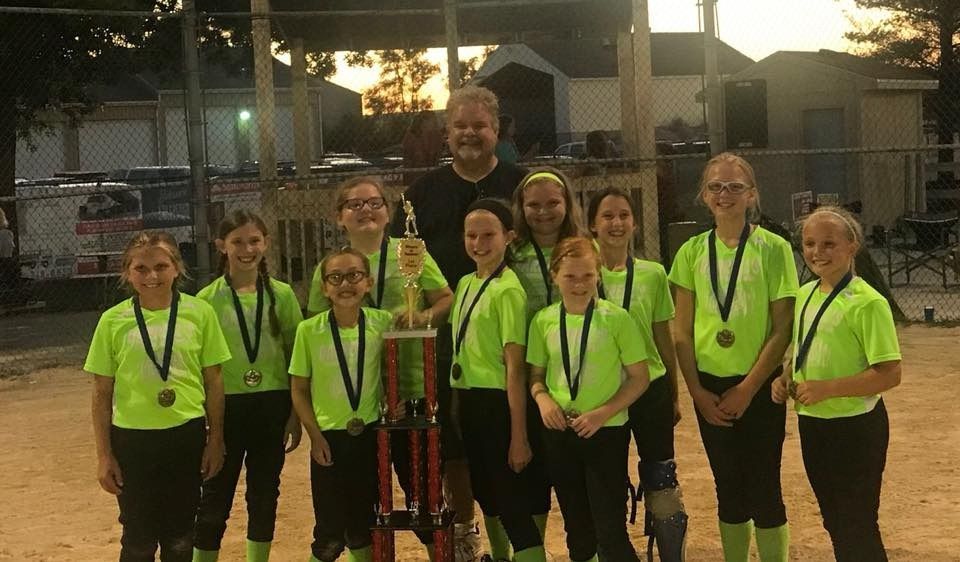 Softball team in neon green shirts and medals, holding a trophy on a dirt field at dusk.
