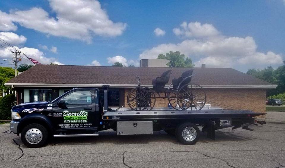 A black tow truck with an antique horse-drawn buggy on its flatbed, parked in front of a building.