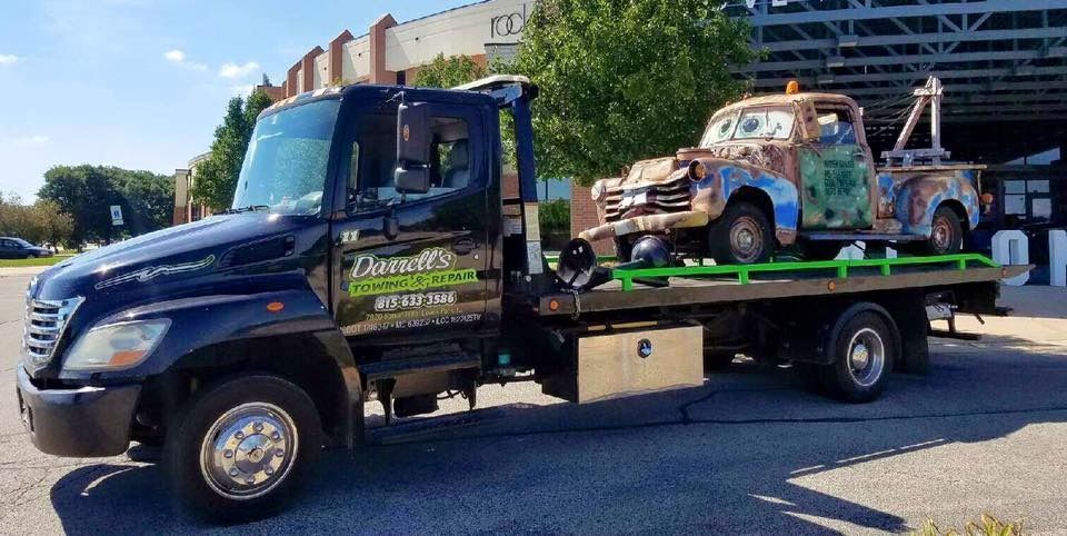 Tow truck carrying a rusty Mater from Cars, parked on pavement.