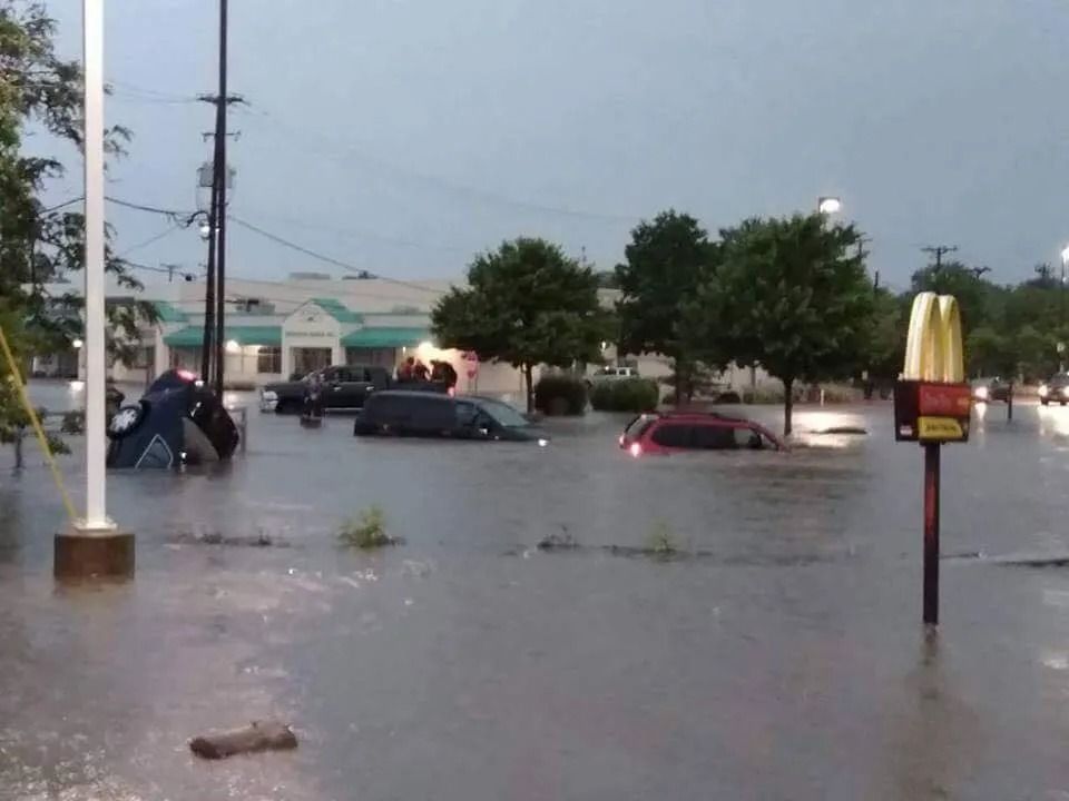 Flooded parking lot with submerged cars, McDonald's sign visible, gray sky, trees, and buildings.