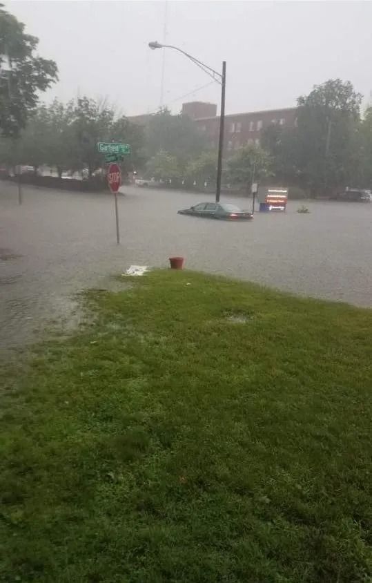 Flooded street with submerged car, sign, and building in the background; green grass in foreground.