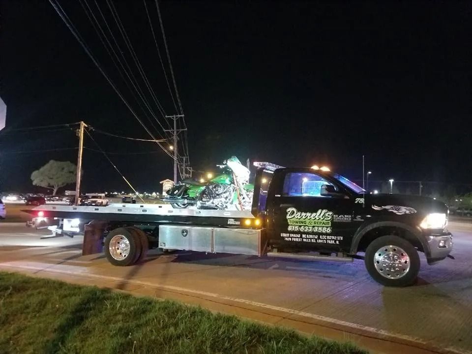 A black tow truck with a damaged green motorcycle on its flatbed at night.