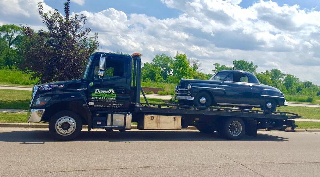 Black tow truck carrying a dark car on a sunny day.