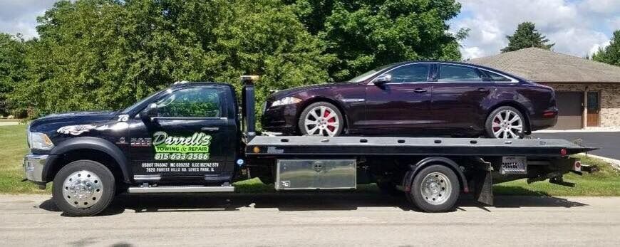 Tow truck carrying a maroon Jaguar sedan. Dark truck with business logo, parked on a road. Green trees in background.