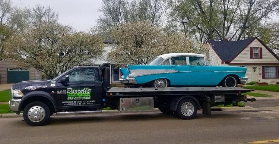 Black tow truck carrying a turquoise classic car on a residential street.