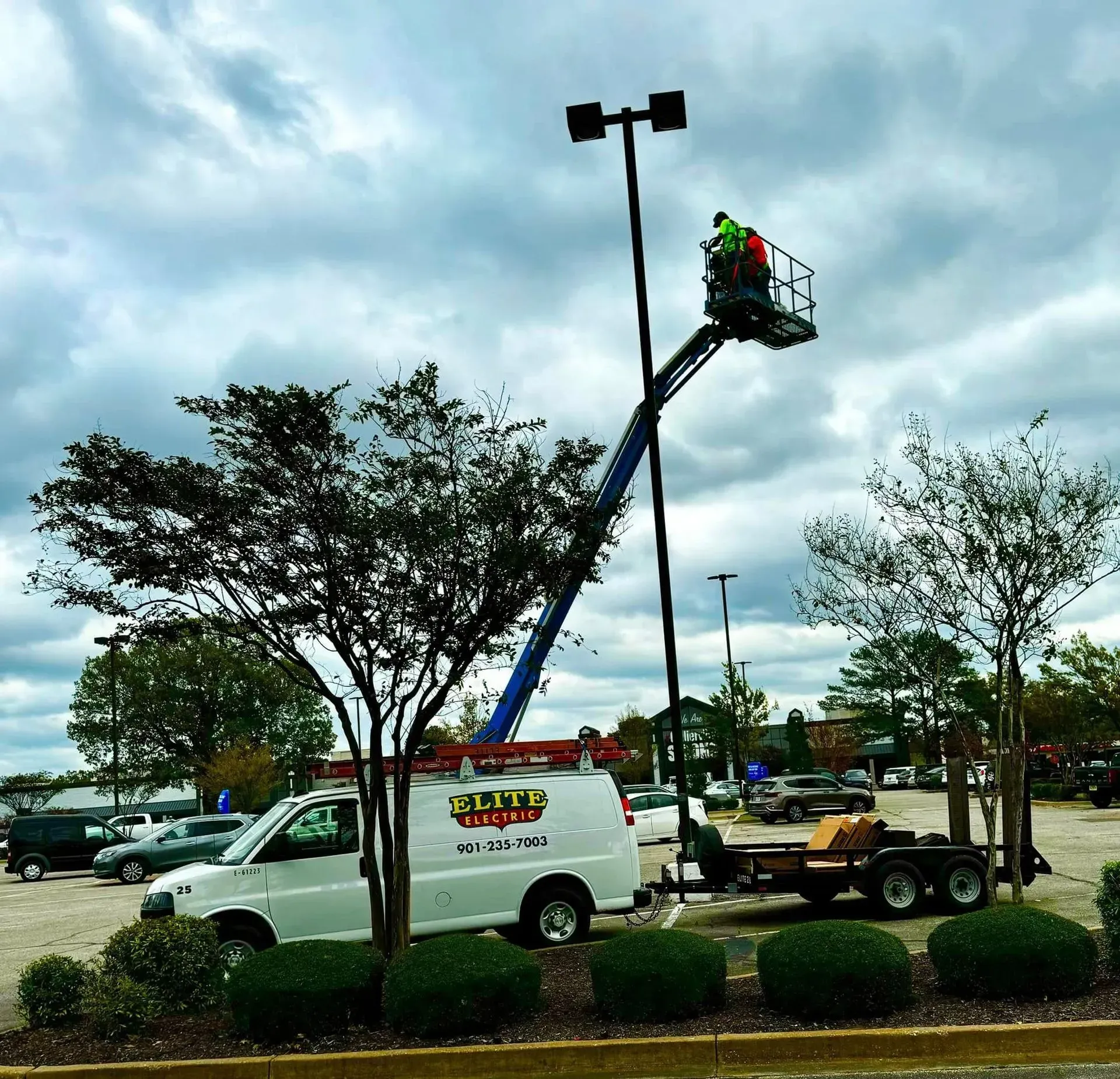 Workers on a lift fixing a parking lot light under a cloudy sky. A white van is parked nearby.