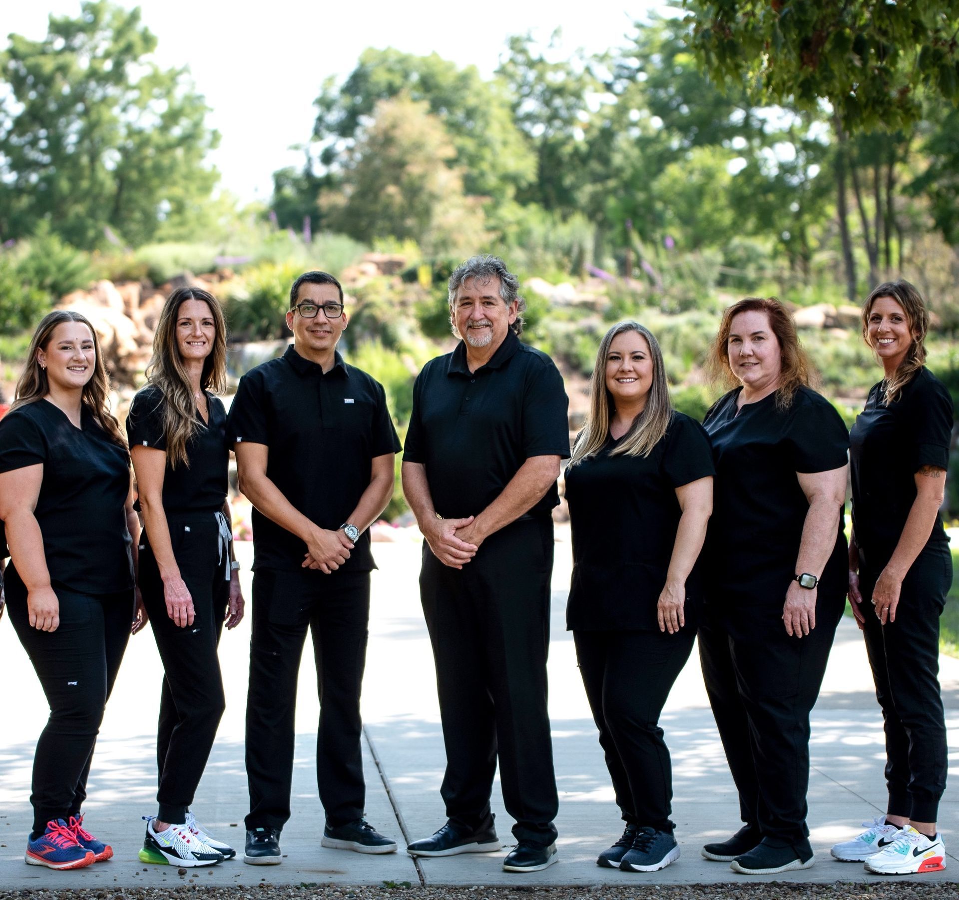 Seven people in black clothing standing on grass in front of trees, arms around each other, smiling.