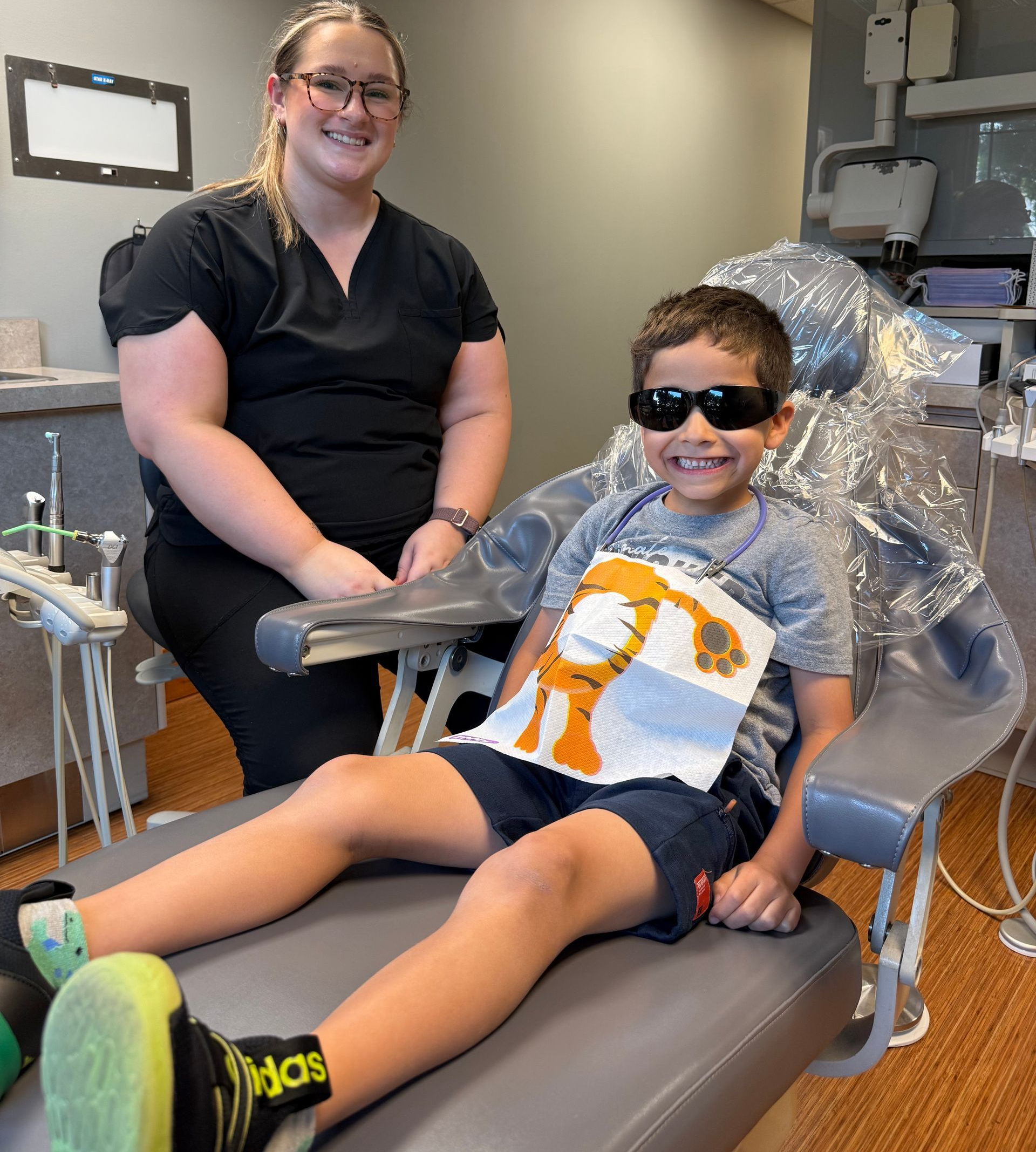 A smiling child in sunglasses at a dentist, with a dental assistant smiling beside him.