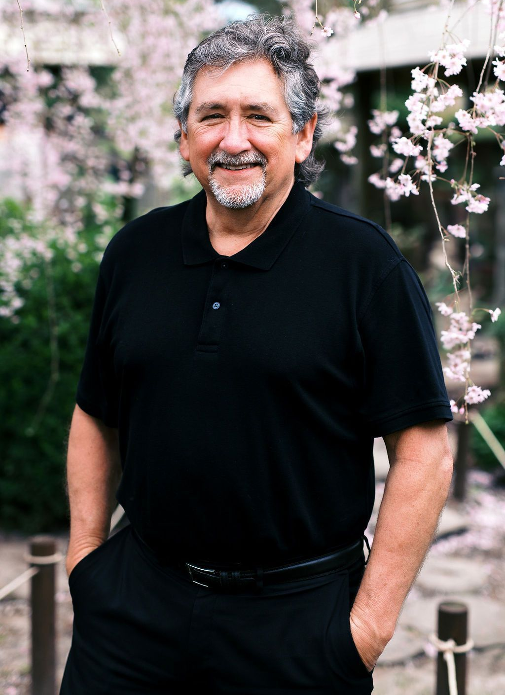 Man with graying hair smiles, hands in pockets, black shirt, blossoms behind him.