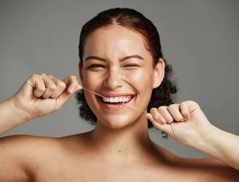 A person winking and smiling while flossing their teeth against a plain gray background.