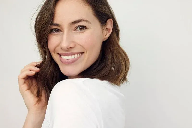 A person with shoulder-length brown hair smiling at the camera against a plain, light gray background.