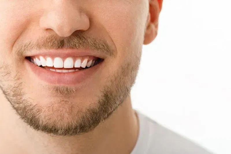Close-up of a person with a short beard, smiling to show bright white teeth, against a plain white background.