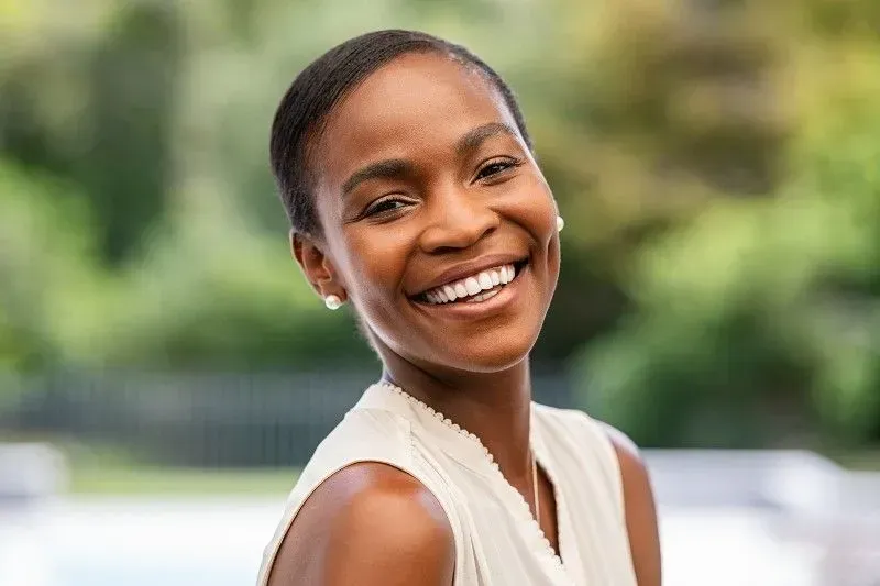 A person with short hair smiling brightly at the camera, wearing a light-colored top with a soft, out-of-focus background.