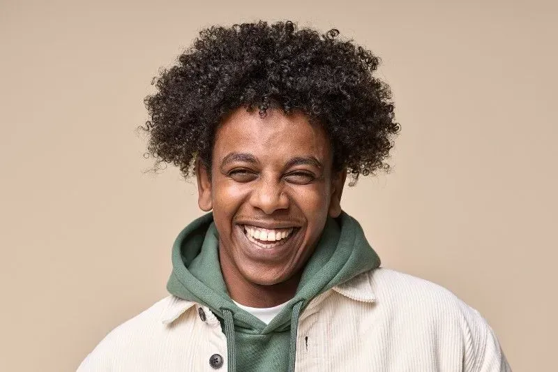 A person with curly dark hair laughing, wearing a green hooded sweatshirt under a cream jacket against a beige background.