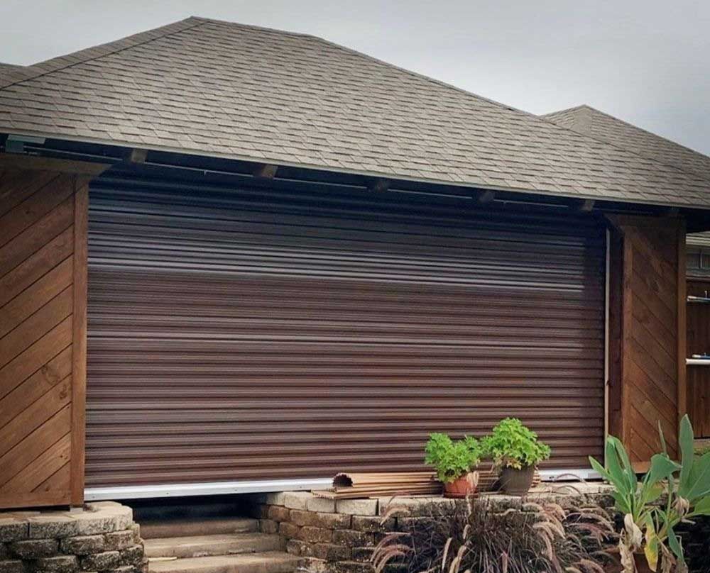 Brown roll-up door on a building with a brown roof and wooden accents, with potted plants in front.