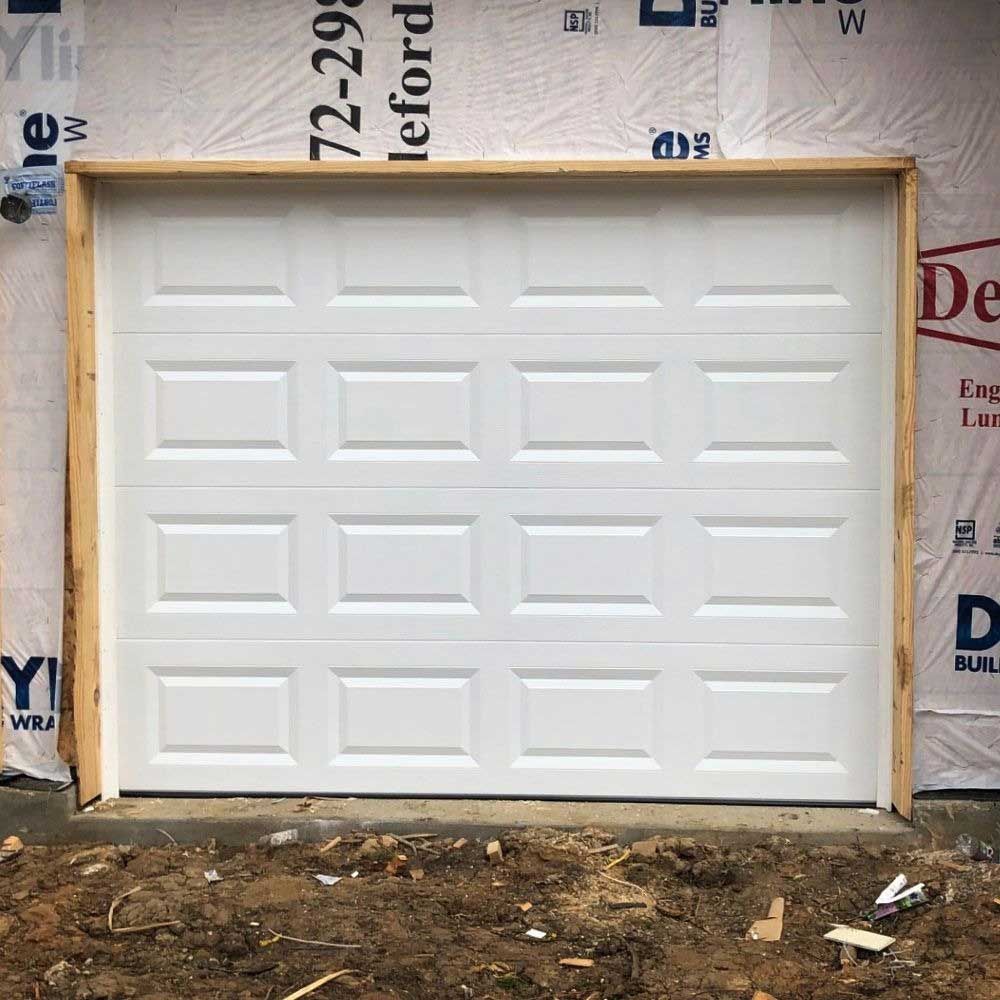 White garage door in a wooden frame, installed in a building under construction, with dirt in front.