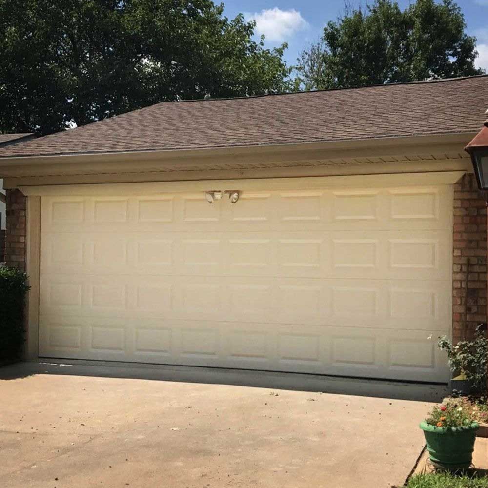 Cream-colored garage door on a brown-roofed house with brick accents, trees in the background, sunny day.