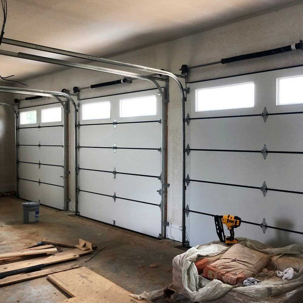 Three white garage doors with windows, inside an unfinished garage.
