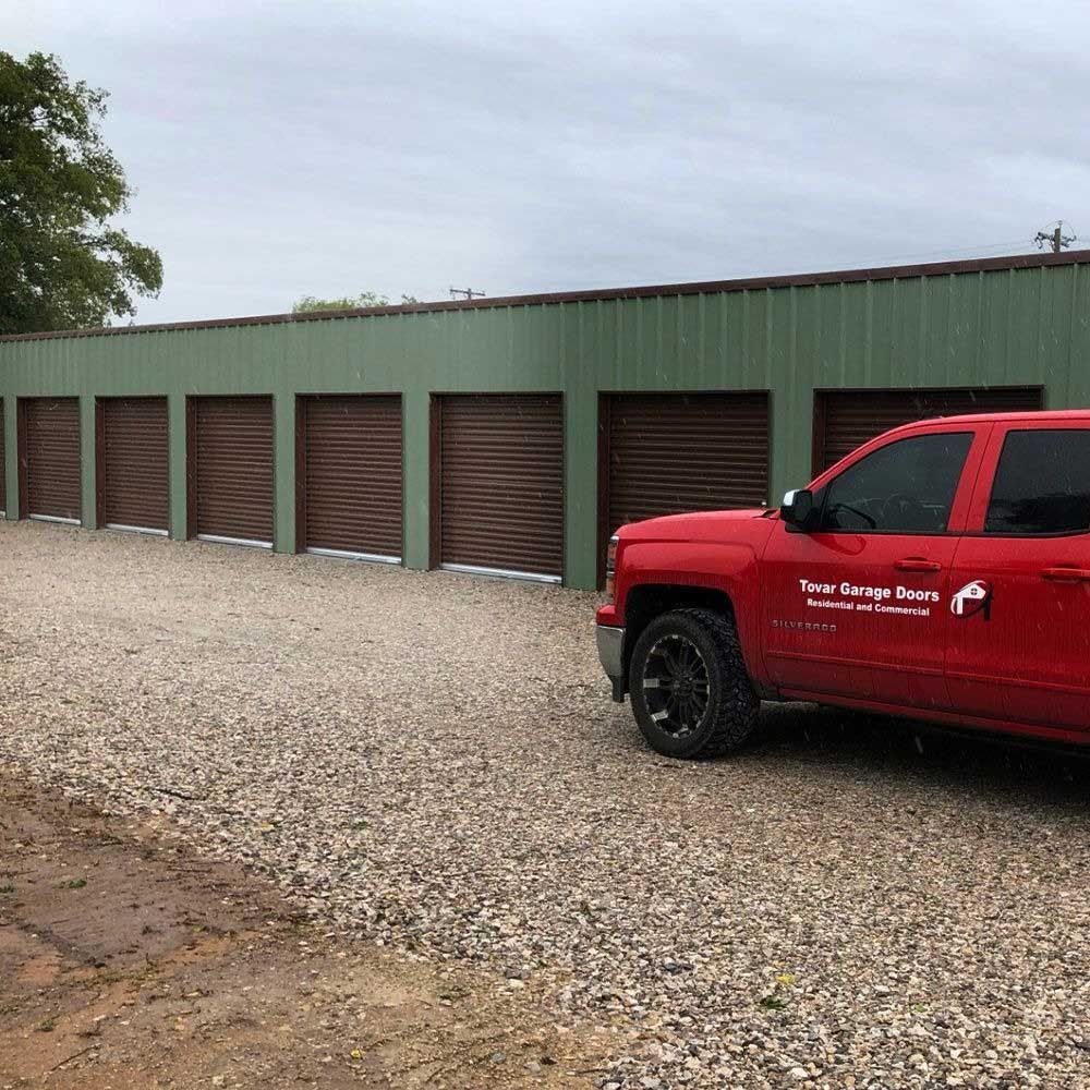 Red truck in front of brown storage unit doors, green building, gravel lot.