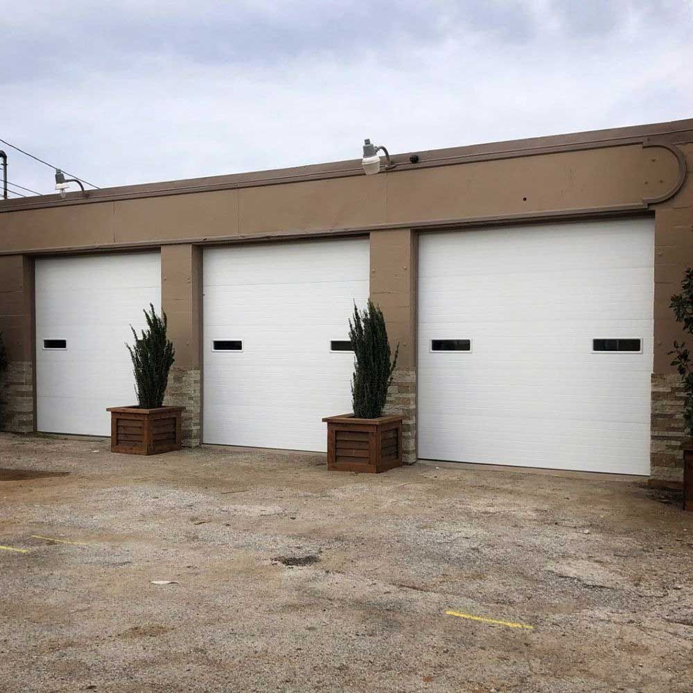 Three white garage doors with black accents, surrounded by tan building and potted plants on a gravel lot.