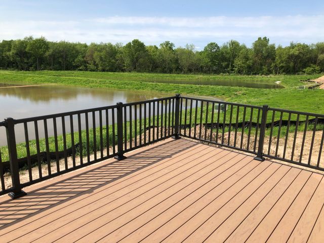 Wooden deck with black railing overlooking a pond and green landscape on a sunny day.