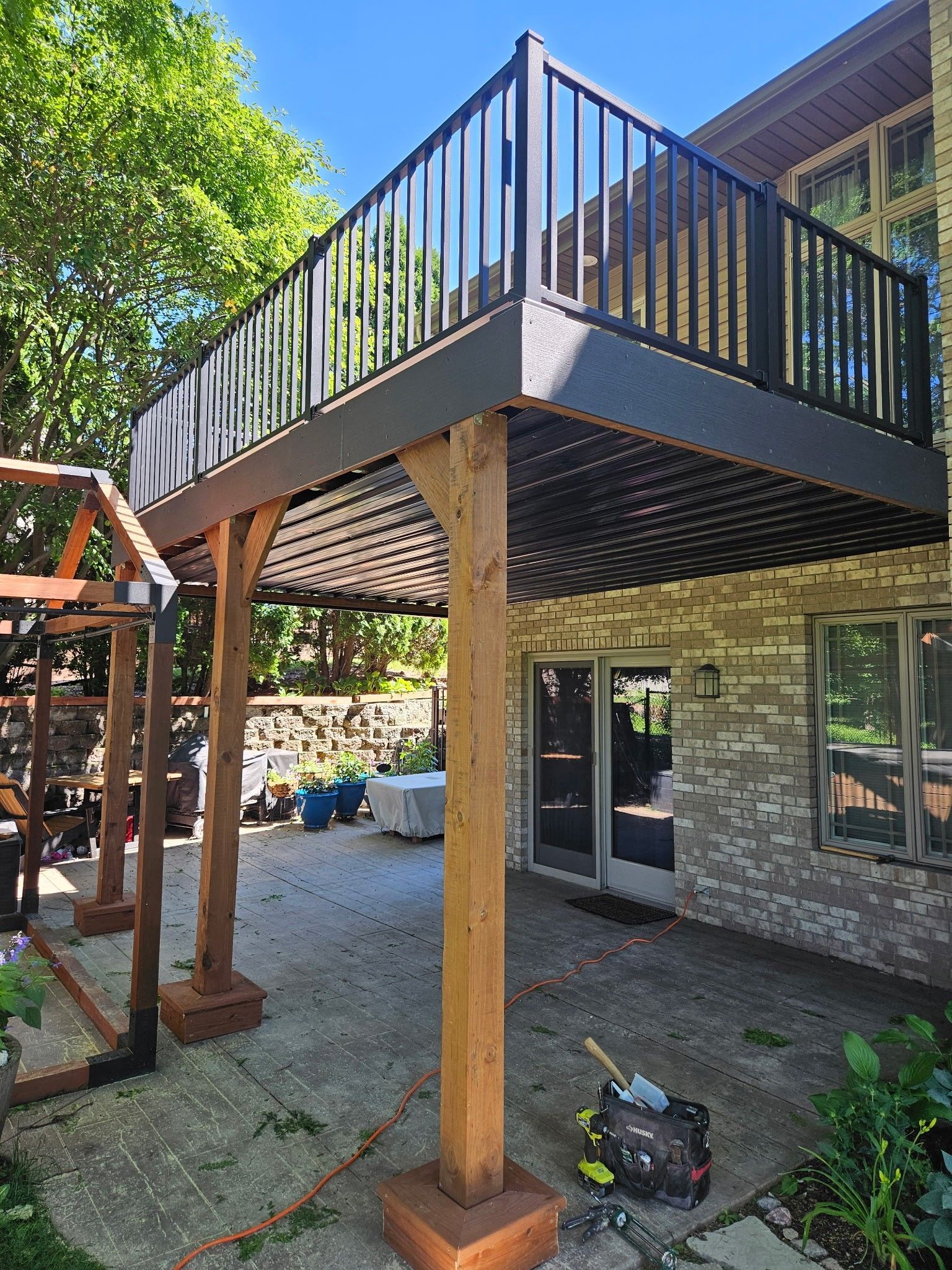 Backyard deck on wooden posts with black railing above a patio and garden area