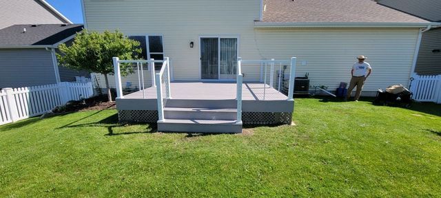Backyard with a deck, green grass, and a white fence. Person stands near some equipment.