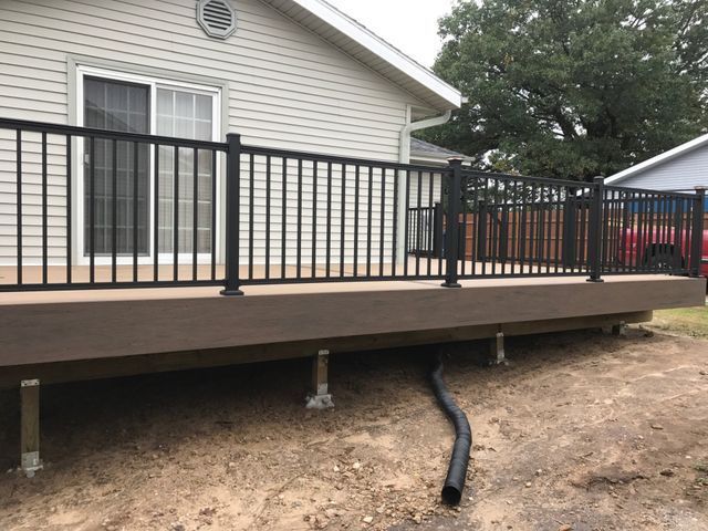 Brown deck with black railing attached to a light-colored house. Drainage pipe visible.