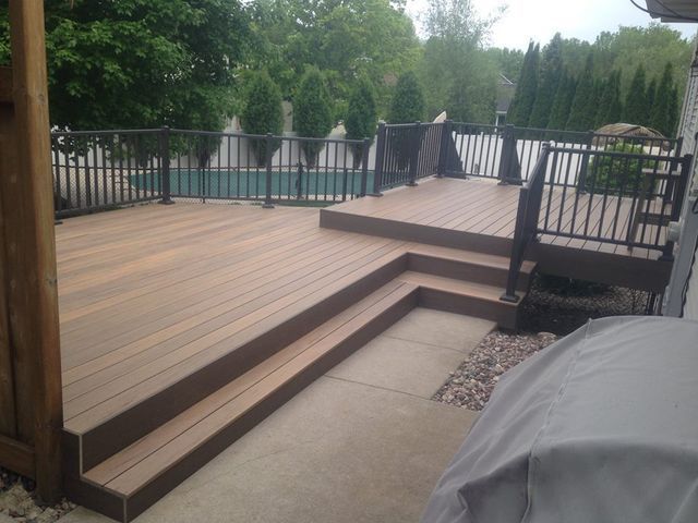Wooden deck with steps, black railing, overlooking a pool and trees. Concrete patio below.