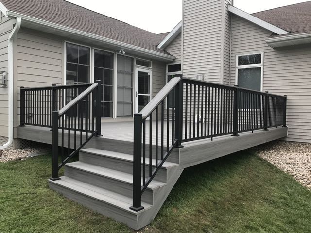 Gray composite deck with black railing and steps attached to a beige house, on a grassy lawn.