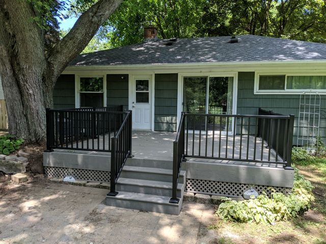 Backyard deck with black railings, steps, and sliding glass doors. Green siding, a large tree, and foliage.