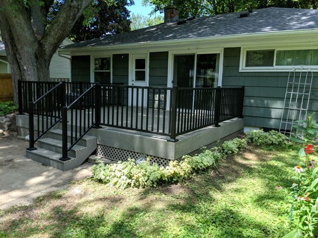 Black railed deck with stairs, attached to a green house with sliding glass doors.