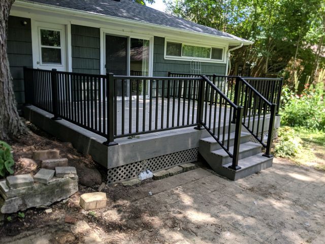 Black railing surrounds a raised concrete deck with stairs leading down to a concrete patio, adjacent to a house.