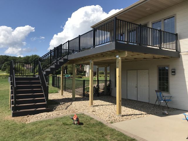 Multi-level deck with black railing and stairs, supported by wooden posts, next to a house with pebble ground.