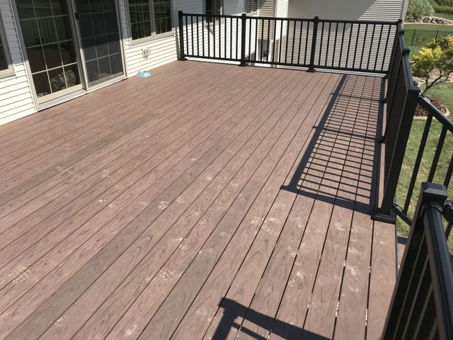Brown composite deck with black railing and light-colored siding. Sunny outdoor setting.