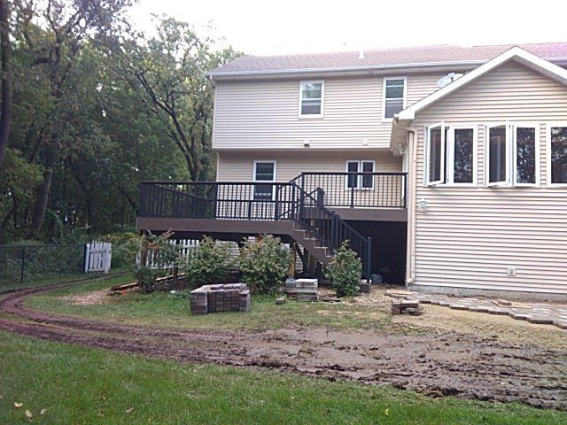 Back of a two-story beige house with a brown deck and steps. Muddy yard with a few bushes and a small fence.