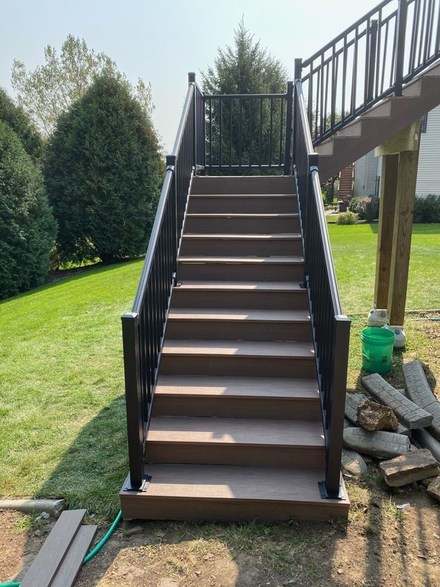 Outdoor wooden stairs with black railing leading up to a deck. Green grass surrounds the stairs.