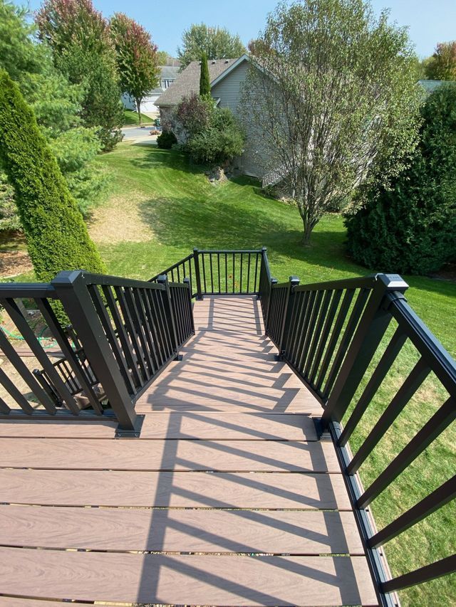 Brown deck with black railing and steps leading down to grassy yard with houses in background.