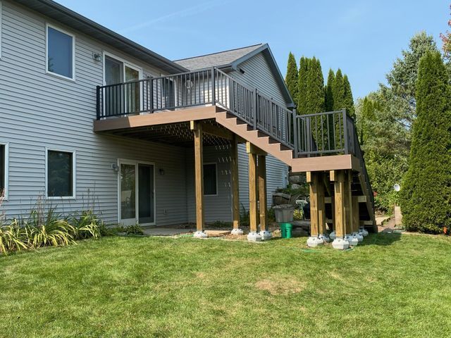 Backyard view of a two-story deck with stairs, supported by wooden posts, next to a gray house, on a sunny day.