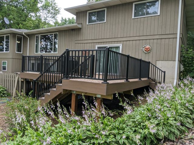 Back of a house with a deck and black railings. Tan siding and stairs. Landscaping with white flowers in front.