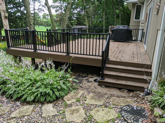A wooden deck with black railings, steps, and a grill, surrounded by greenery and stone pathways.