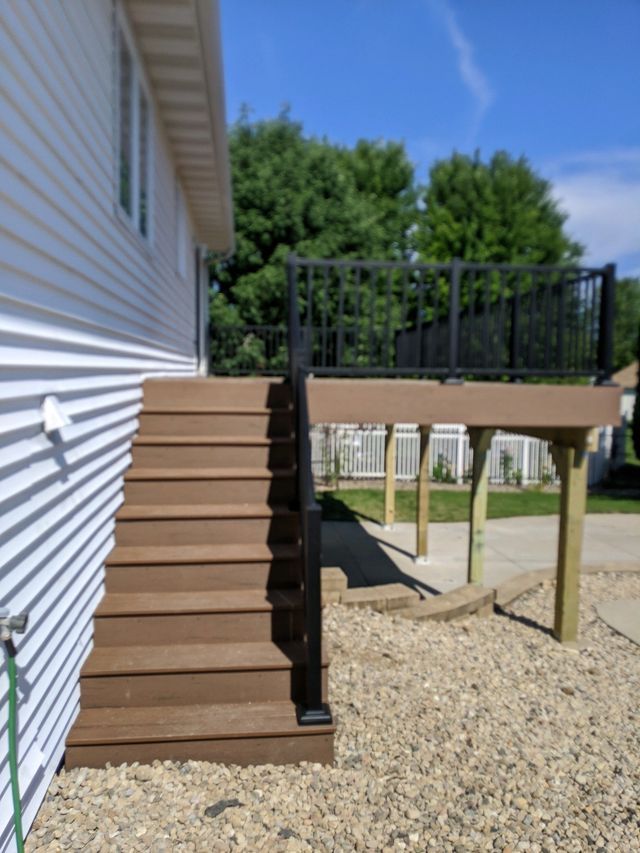Brown wooden stairs and deck with black railing attached to a white house, gravel ground.