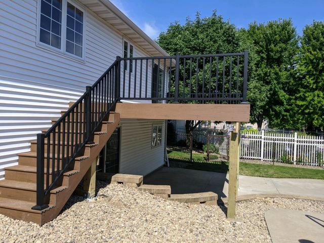 Brown deck and stairs with black railing attached to a white house, gravel ground, sunny day.
