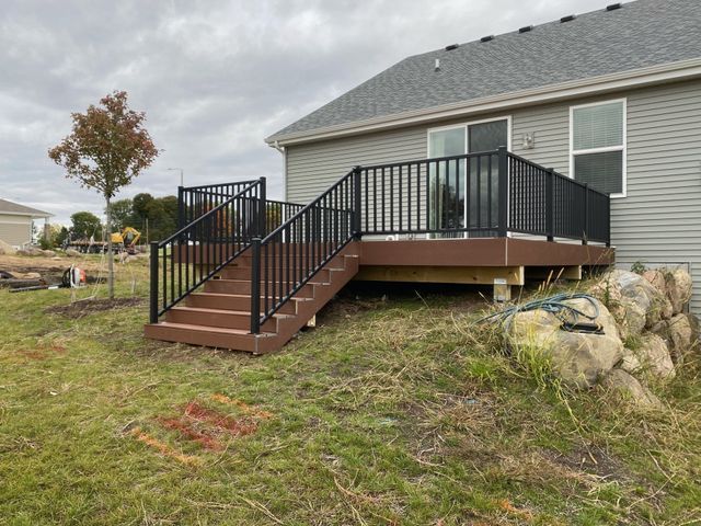 A wooden deck with black railings and stairs on a grassy hill next to a house with gray siding.