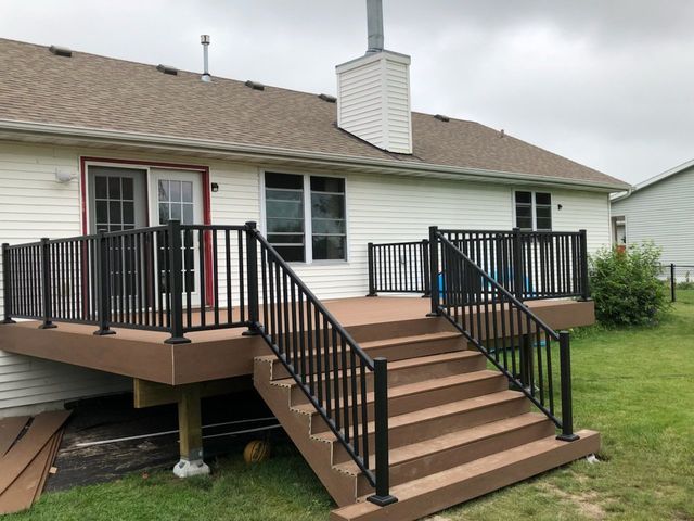 Brown composite deck with black railings and stairs; attached to a beige house, with green grass in yard.
