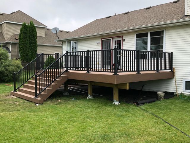 Brown deck with black railing and stairs attached to a light-colored house. Green grass.