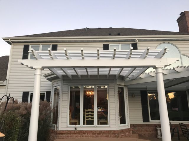 White pergola attached to a light-colored house with a dark roof.