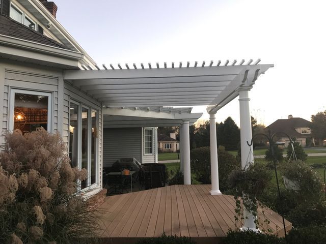 White pergola over a wooden deck attached to a house with large windows.