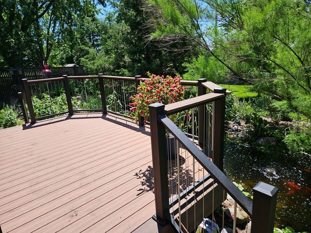 Wooden deck with dark railing and stainless steel cables overlooking a pond with fish and lush green trees.
